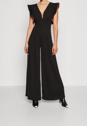 Woman wearing black wide-leg jumpsuit with deep V neckline and ruffled cap sleeves, paired with black ankle boots with white toe caps.