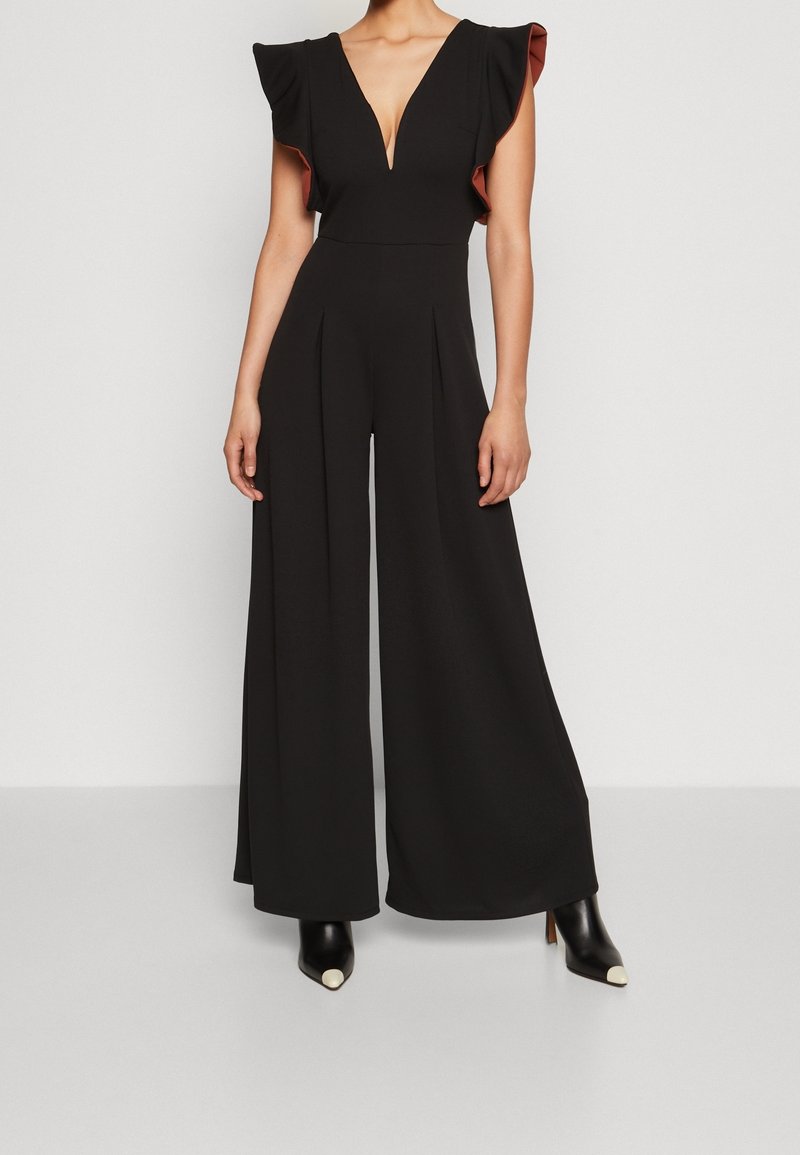 Mujer vestida con un mono negro de pierna ancha con escote en V profundo y mangas de volante, combinado con botas de tobillo negras con punteras blancas.