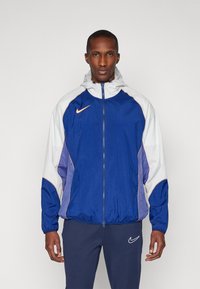 Veste coupe-vent en matériau synthétique, avec un design en blocs de couleurs bleues et blanches, fermeture zippée et poignets élastiques. Logo Nike sur la poitrine.