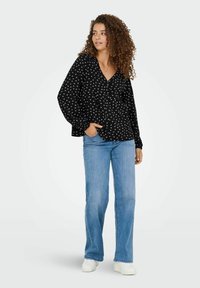 Zwarte blouse met knoopjes, witte stippen, lange pofmouwen en een v-hals, gecombineerd met lichtblauwe flare jeans en witte sneakers.