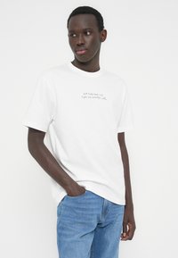 Νεαρός με σκουρόχρωμη επιδερμίδα που φοράει λευκό t-shirt με κείμενο και ανοιχτό μπλε τζιν παντελόνι, στέκεται με το ένα χέρι στην τσέπη μπροστά σε απλό φόντο.