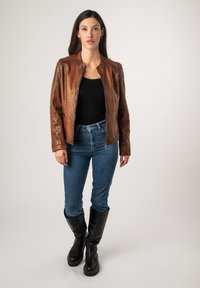 Bruine leren jas met ritssluiting, ronde hals en lange mouwen. Draagt over een zwarte top, gecombineerd met blauwe jeans en hoge zwarte laarzen.
