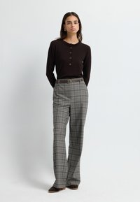 Top marrón de mangas largas con un cuello fruncido y detalle de botones, combinado con pantalones de cuadros gris de talle alto y zapatos marrones.