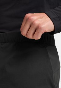 Pantalons noirs à taille élastique avec une texture lisse, affichant un design ajusté. La main saisit la taille pour un ajustement.