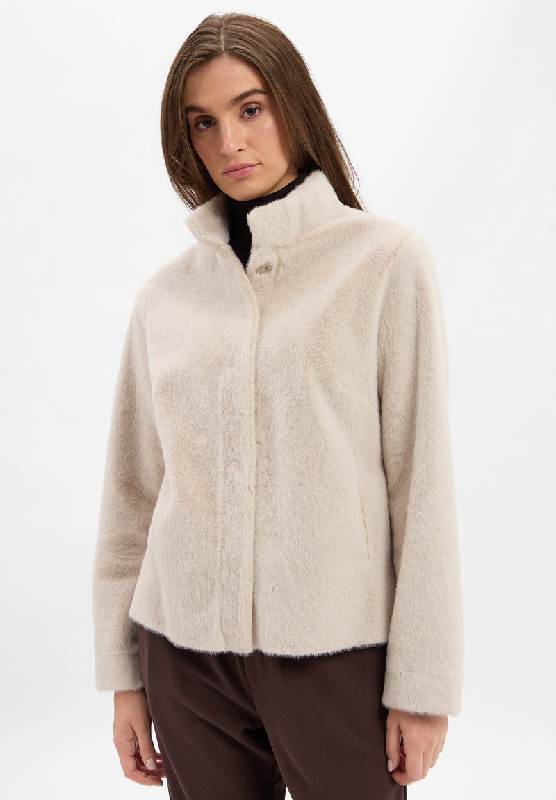 Beige neplederen bontjas met een hoge kraag, sluiting met knopen aan de voorkant en zijzakken. Zachte textuur en relaxte pasvorm, gecombineerd met een bruine broek.