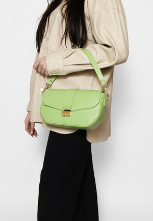 Donna con giacca beige e pantaloni neri, con una borsa a tracolla in pelle verde chiaro con chiusura dorata.