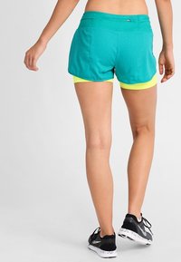 Pantalones cortos de correr color teal con un diseño texturizado, que cuentan con un pantalón interior amarillo en contraste, combinados con zapatillas de correr negras.