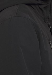 Černá kapucí s hladkým, matným povrchem, se zipovými kapsami a nastavitelnými šňůrkami na kapuci. Žádné viditelné vzory.