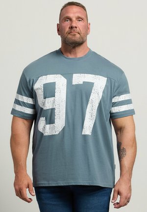 Hombre adulto con pelo corto y barba, vestido con una camisa azul grisácea con el número blanco grande 97 y franjas blancas en las mangas, de pie contra un fondo liso.