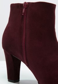 Bordeaux ruskindsankelstøvle med lynlås i siden, med en rundet tå og en solid hæl, designet til en tætsiddende silhuet.