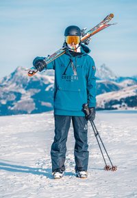 Skijder in blauwe jas, zwarte helm en oranje skibril houdt ski's over de schouder en skistokken vast, staande in de sneeuw met bergen op de achtergrond.