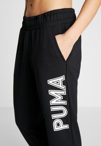 Zwarte sportbroeken met een elastische tailleband, zijzak en groot wit "PUMA"-logo op het linkerbeen gedrukt.