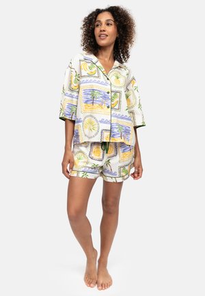 Frau trägt kurzärmliges Hemd mit Knöpfen und passende Shorts mit bunten tropischen Früchten und Strandmotiven, steht barfuß.