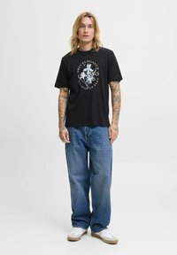 Sort bomuld t-shirt med et centralt hvidt grafisk print, kombineret med løse denim jeans og hvide sneakers med brune accenter.