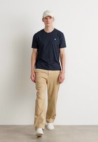 Navy kortærmet t-shirt, beige bukser og hvide sneakers. En lys kasket med tekst fuldender outfittet. Model står foran en neutral baggrund.