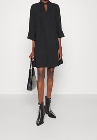 Robe décontractée noire avec un col montant, des manches trois-quarts et un détail noué. Portée avec des bottines noires pointues et un sac à main noir texturé.