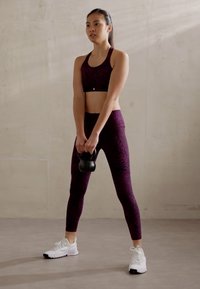 Purpurno vzorčasta športna modrček in legice, drži črno kettlebell, obuta v bele športne čevlje, stoji na sivi tleh s betonsko ozadje.