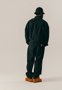 Mustat denim-takki ja housut, teksturoitu kangas, takataskussa ruskea merkki; yhdistetty oransseihin työsaappaisiin ja mustaan ämpärihattuun.