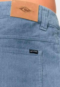 Pantalones cortos de pana azul con textura de ribetes verticales, parche de cuero marrón con logo y una pequeña etiqueta negra en la costura del bolsillo.