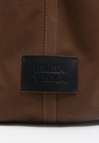 Brūnas auduma soma ar melnu īstas ādas etiķeti, uz kuras ir iepresēts "BIMBA Y LOLA", apvilkta ar kontrastējošiem šuvēm un teksturētu materiālu.