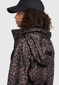 Giacca impermeabile marrone scuro con motivo astratto rosa, dotata di un colletto alto, cappuccio regolabile e tasche con zip. Cappellino nero indossato.