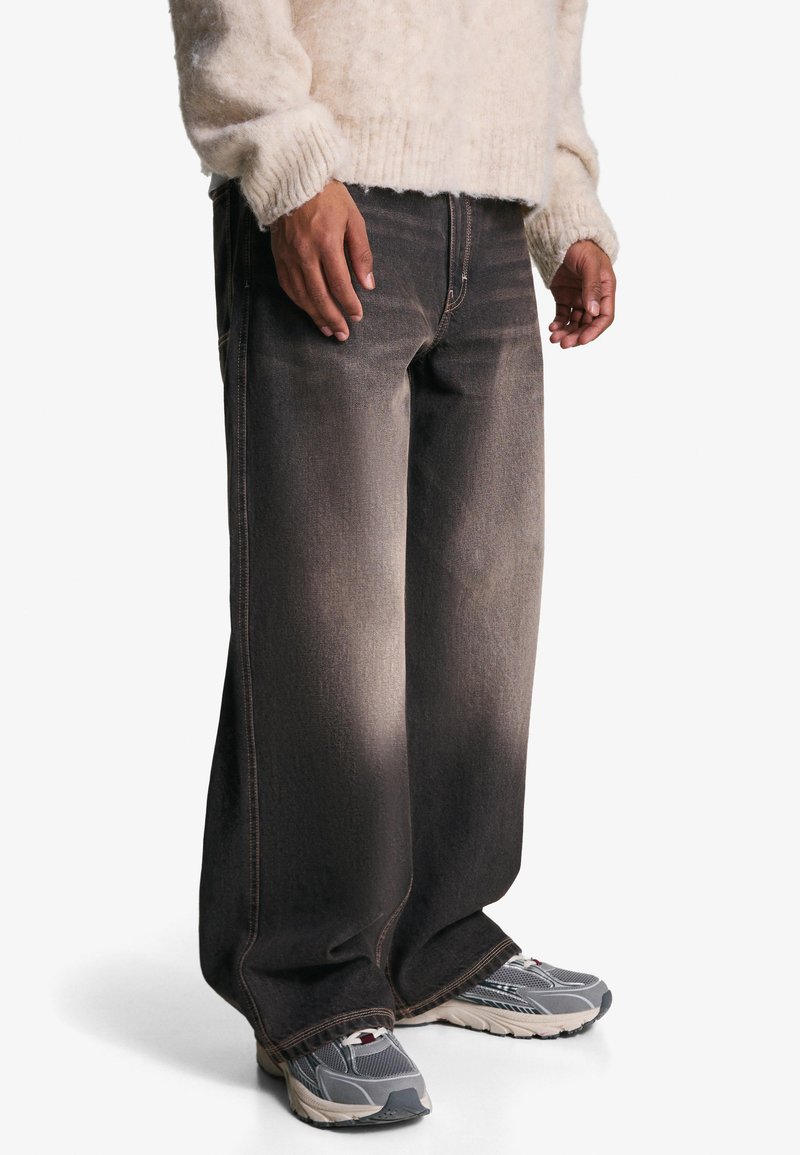 Weite, dunkelgraue Jeans aus Denim mit einem ausgewaschenen Finish, kombiniert mit grauen Sneakers, die kontrastierende Nähte aufweisen und eine lockere Passform bieten.