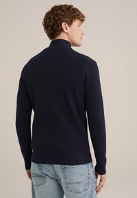 Marineblauer Strickpullover mit hohem Kragen, gerippten Bündchen und Saum. Hergestellt aus strukturiertem Material, getragen mit hellblauen Jeans.