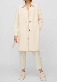 Abrigo de mezcla de lana color beige claro con cuello con solapa, grandes bolsillos delanteros y botones oscuros. Combinado con pantalones crema claros y zapatillas deportivas gruesas.