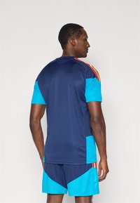 Vīrietis stāv ar muguru pret mums, valkājot tumši zilu un gaiši zilu sporta apģērbu ar sarkanām un dzeltens svītrām uz pleciem un sāniem.