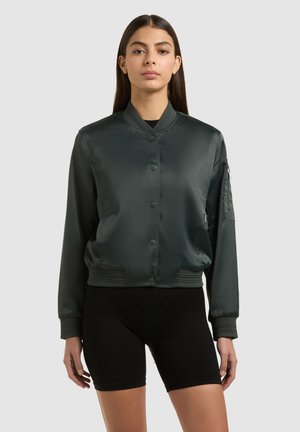 Kobieta ubrana w ciemnozieloną satynową kurtkę typu bomber z zatrzaskami i czarne spodenki rowerowe, stojąca na tle jednolitego jasnego tła.