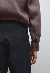 Zwarte broek met een getailleerd ontwerp, voorzien van achterzakken. Gedragen met een losse, bordeauxrode, geknoopte blouse met lange mouwen. Effen textuur.