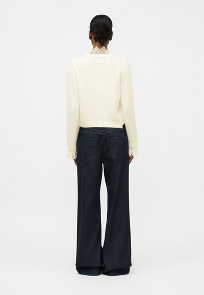 Γυναίκα με σκούρα μαλλιά σε κότσο, φορώντας εκρού πουλόβερ με δαντελένια μανίκια και γιακά, συνδυασμένο με σκούρο τζιν παντελόνι φαρδιάς γραμμής, στέκεται με την πλάτη στραμμένη.