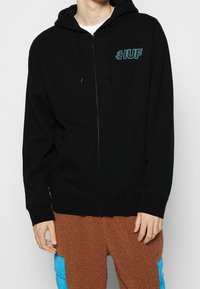 Felpa nera con zip e tasca frontale, caratterizzata da un logo "HUF" turchese sul lato sinistro del petto. Realizzata in materiale morbido con cappuccio regolabile.