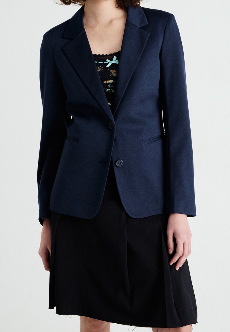 Femme portant un blazer bleu marine sur un haut fleuri avec un nœud vert sarcelle, assorti à une jupe noire, sur un fond clair uni.
