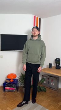 Olivno zelena jopa s kengurujim žepom, v kombinaciji s črnimi hlačami in temnimi loaferji. Dodatki vključujejo črno kapo in tetovaže, vidne na rokah.