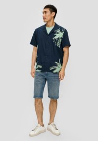 Korte mouwen marineblauw shirt met een lichtgroen palmbomenprint, knoopsluiting, gecombineerd met lichtblauwe denim shorts en witte sneakers.