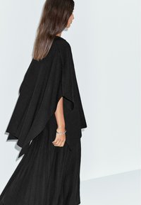 Poncho noir au design fluide et oversize. Confectionné dans un tissu doux, avec un col montant et des manches drapées, associé à une jupe plissée noire.