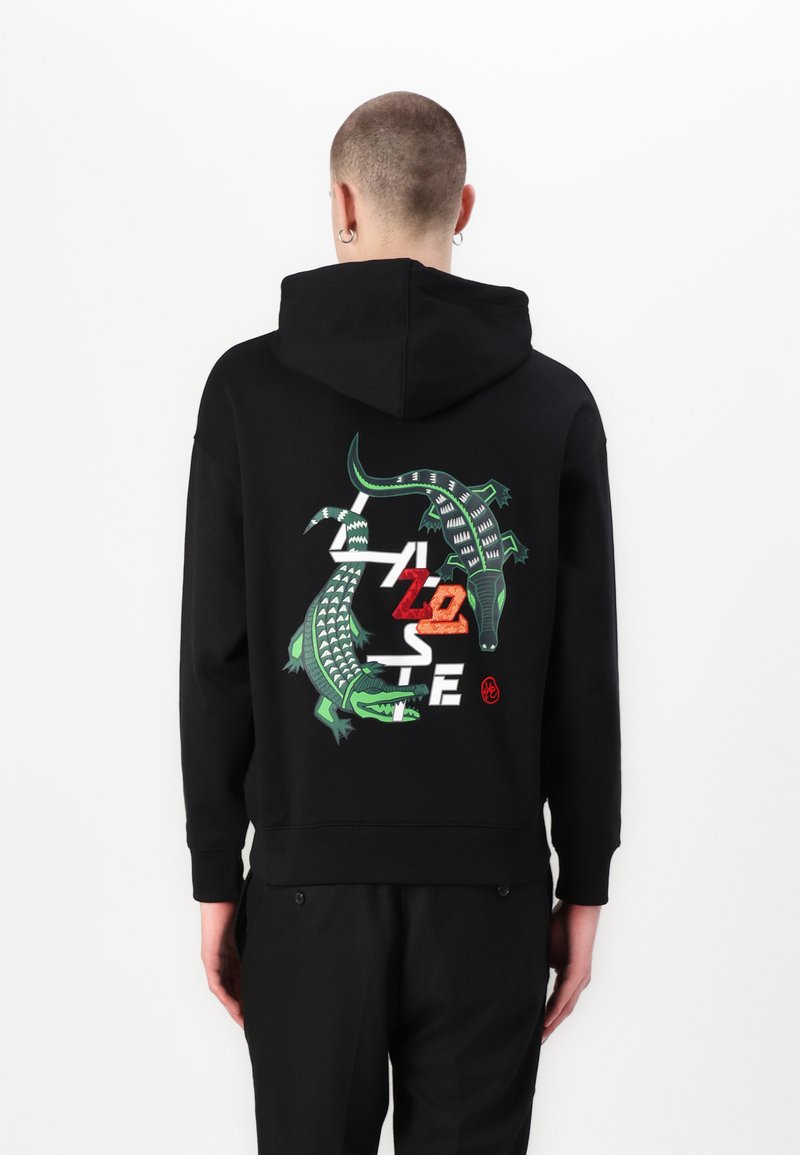 Fekete kapucnis pulóver zöld krokodil grafikával, piros és fehér felirattal, valamint puha tapintással. Kapucnis kivitel, bordázott mandzsettákkal és alsó résszel.