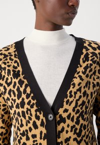 Bež kardigan s leopardjim vzorcem in črnimi detajli, s krojem v obliki črke V ter zapiranjem na en črn gumb. Nosi se čez belo polo majico.