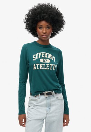 Grünes Langarm-T-Shirt mit aufgedrucktem "SUPERDRY ATHLETIC 87" in Weiß. Mit Rundhalsausschnitt und kombiniert mit hellblauen Jeans.