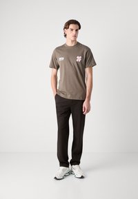 Olive green cotton t-shirt featuring a pink floral graphic and text on the left chest, paired with black trousers and white sneakers.