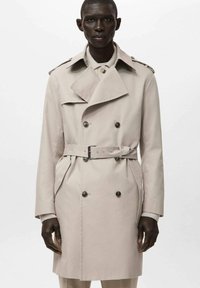 Beżowy dwurzędowy płaszcz trencz z paskiem w talii, dużym kołnierzem, pagonami na ramionach i bocznymi kieszeniami. Gładka, wypolerowana faktura tkaniny.