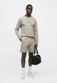 Svetlo bež majica z napisom »EA7 Emporio Armani«, ustrezajoče kratke hlače, bele nogavice in svetli športni copati. Drži črno torbo.