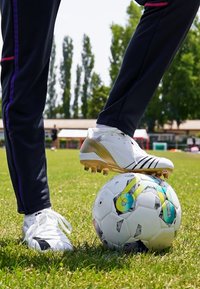 Biele futbalové kopačky so zlatými akcentmi ležia na vzorovanej futbalovej lopte, obklopené zelenou trávou a jasnou oblohou. Tmavé športové nohavice dopĺňajú celkový vzhľad.