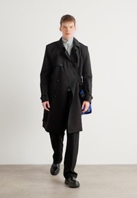 Black trench coat with double-breasted buttons, worn over a gray top and black trousers. Blue bag and chunky black shoes complete the look.