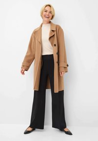 Beige Wollmantel mit großem Kragen und vier schwarzen Knöpfen, kombiniert mit einem hellen Langarmoberteil und schwarzen weiten Hosen sowie Schuhen.