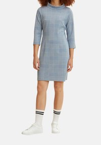 Vestido de cuadros azul con cuello de embudo y mangas tres cuartos. Hecho de una tela texturizada. Combinado con zapatillas blancas y calcetas de rayas hasta la rodilla.