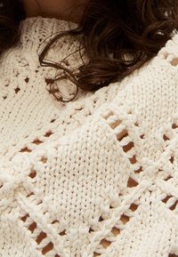 Pull en maille crème avec motifs ajourés, présentant diverses textures et points, affichant un design ample et confortable.