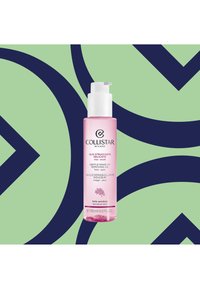 Transparante fles met roze makeupremoving olie en een witte pomp. Etiket bevat de productnaam en meertalige tekst. Achtergrond heeft groene en marineblauwe patronen.
