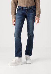 Person in blauen geraden Jeans, weißen auffälligen Sneakers und einem beigen Strickpullover, der auf einem weißen Boden vor einem schlichten Hintergrund steht.